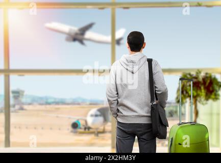 Il turista maschile calmo è in piedi in aeroporto e guardando il volo aereo attraverso la finestra. Ha in mano biglietti e valigia. Tramonto Foto Stock