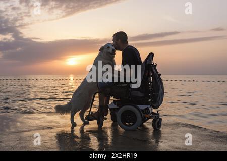 Uomo con disabilità, utente su sedia a rotelle elettrica accarezzare il cane e godersi un tramonto sulla spiaggia. Concetto di amicizia tra persone e animali. Foto Stock