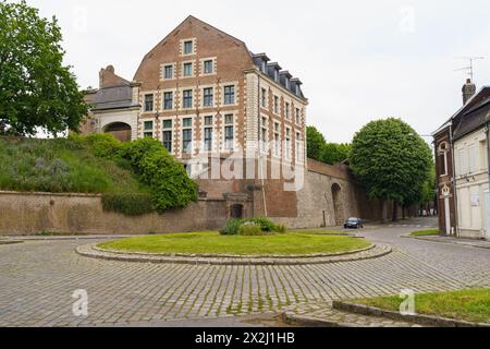 Cambrai, Francia - 21 maggio 2023: Un consistente edificio in mattoni si erge alto su un campo verde vivace sotto un cielo limpido, mostrando il contrasto tra m Foto Stock