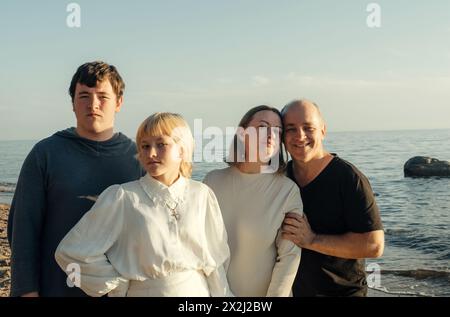 Una famiglia di quattro persone si trova vicino al mare, abbracciandosi l'un l'altro mentre si crogiola nel soffice bagliore del sole che tramonta. Foto Stock