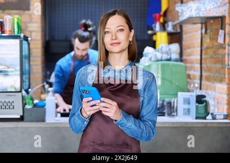 Giovane lavoratrice di servizio in grembiule con smartphone nel ristorante e nella caffetteria Foto Stock