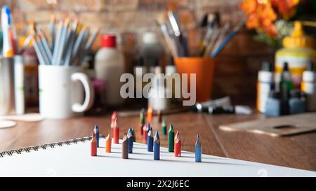 Piccole matite colorate su carta bianca su un tavolo di legno con materiali artistici sullo sfondo fuori fuoco Foto Stock