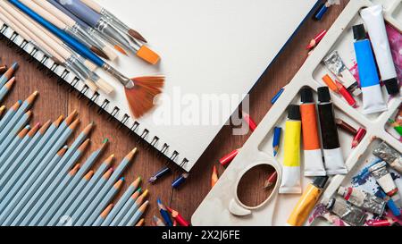 Vista dall'alto del tavolo in legno con matite, tubi di vernice, materiali d'arte, varietà di pennelli su carta bianca Foto Stock