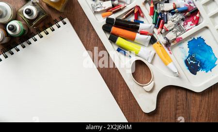 Vista dall'alto del tavolo in legno con matite, tubi di vernice, materiali d'arte, flaconi d'inchiostro, varietà di pennelli su carta bianca Foto Stock