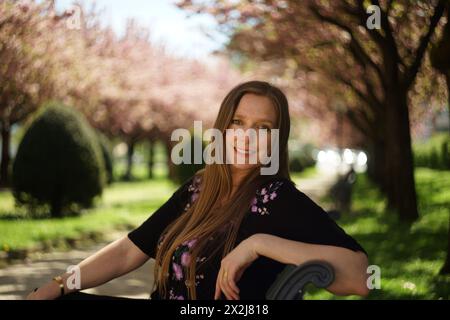 Donna europea siede su una panchina del parco sullo sfondo di un vicolo di ciliegi ornamentali in piena fioritura con delicati fiori rosa, lussureggiante verde gr Foto Stock