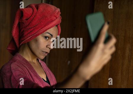 Giovane uomo transgender (26) con accappatoio, asciugamano che copre la testa e cellulare in mano facendo un autoritratto. Concetto transgender Foto Stock