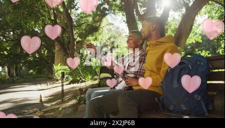 Immagine di cuori rosa che fluttuano sopra una coppia gay maschio felice e diversificata con caffè che parlano nel parco Foto Stock