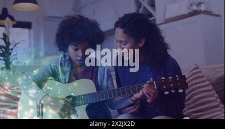 Immagine di macchie luminose su madre birazziale felice con figlio che suona la chitarra Foto Stock