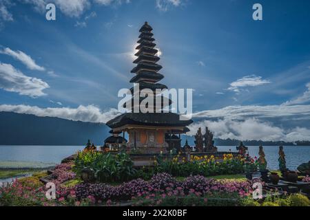 Questa suggestiva immagine cattura un antico tempio con paglia a più livelli, vividamente circondato da fiori colorati e affacciato su un tranquillo lago contro un B. Foto Stock