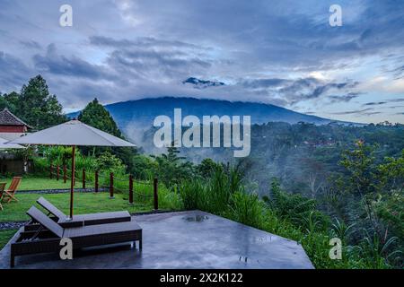 Una serena vista mattutina da un lussuoso patio del resort, caratterizzato da lettini e un ombrellone che si affaccia su un paesaggio montano nebbioso, che incarna tranquillità e Foto Stock