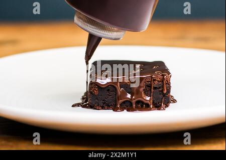 La salsa di cioccolato viene versata sul brownie su un piatto bianco sul tavolo del ristorante in legno Foto Stock