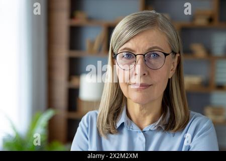 Ritratto di una donna anziana sicura di sé che indossa occhiali e una camicia azzurra, ambientato in un moderno ambiente d'ufficio con uno sfondo di scaffali. Foto Stock