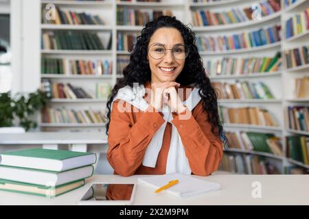 Una giovane donna allegra che indossa occhiali e un abbigliamento casual sorride alla macchina fotografica mentre si siede a un tavolo in una biblioteca circondata da libri. Foto Stock