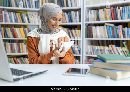 Una giovane donna musulmana, che indossa un hijab, che si impegna nei suoi studi in una biblioteca. Sta scrivendo appunti mentre usa un computer portatile e circondata da libri. Foto Stock