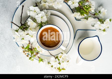 Una vista dall'alto di una tazza di caffè circondata da fiori di primavera bianca e una caraffa per il latte, su uno sfondo bianco Foto Stock