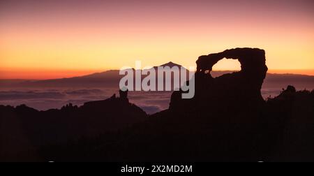 Una splendida vista crepuscolare della silhouette di Roque Nublo sullo sfondo del soffice bagliore del tramonto con il Monte Teide sullo sfondo, sopra una Gran ca coperta di nuvole Foto Stock