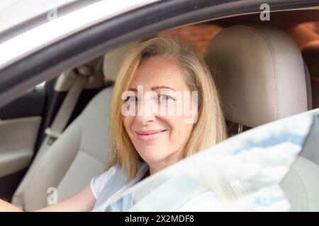 Una donna matura dai capelli biondi è seduta sul sedile di un'auto e sorride davanti alla telecamera attraverso la finestra aperta Foto Stock