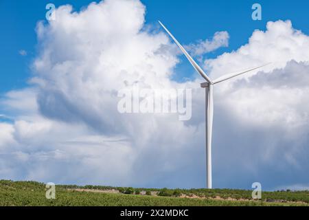 Una turbina eolica solitaria si erge in alto con le sue pale che tagliano attraverso un cielo blu vibrante punteggiato da soffici nuvole bianche, raffigurando la generazione di S. Foto Stock