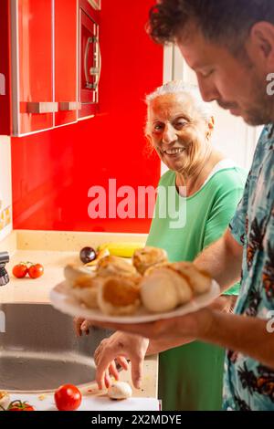 Una donna e un uomo preparano il cibo in una cucina rossa, sorridendo mentre si divertono con la famiglia prima di un barbecue con la nonna. Foto Stock