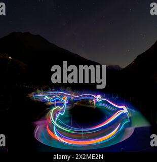 Una scena notturna che cattura le colorate piste luminose create dai ciclisti su una pista ciclabile tortuosa, con montagne e cielo stellato sullo sfondo, durante il summ Foto Stock