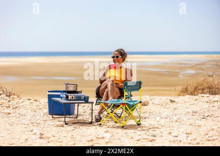 Una donna allegra siede su una sedia da campeggio, sorseggiando un drink, con una stufa portatile accanto a lei durante un viaggio in campeggio nel panoramico sud di Fuerteventura, Foto Stock