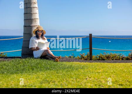 Una tranquilla scena di una donna che si rilassa con un libro accanto all'oceano sotto una palma, godendosi l'ambiente sereno. Foto Stock