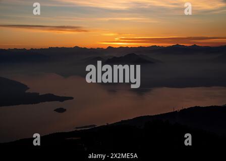 Un tramonto sereno proietta calde sfumature sul Laggo maggiore, con le sagome delle montagne e delle isole che emergono dalla nebbia Foto Stock