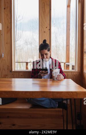 Una giovane donna che indossa una camicia a quadri è infilata nel suo smartphone mentre tiene in mano una tazza, seduta ad un tavolo di legno vicino a una finestra con una vista serena Foto Stock