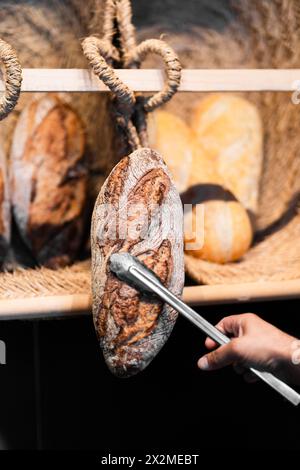 Una mano corta e irriconoscibile che utilizza pinze per raccogliere un pane rustico a lievitazione naturale da una selezione di tonalità calde visualizzata su un ripiano di legno. Foto Stock