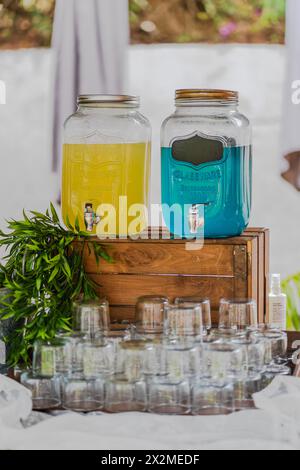Un'elegante postazione per bevande per matrimoni con due grandi distributori di bevande gialle e blu, circondata da bicchieri ordinati Foto Stock