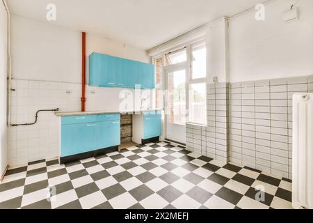 Una cucina vuota e illuminata dal sole presenta armadi blu luminoso, piastrelle bianche e un classico pavimento a scacchi bianco e nero. Foto Stock
