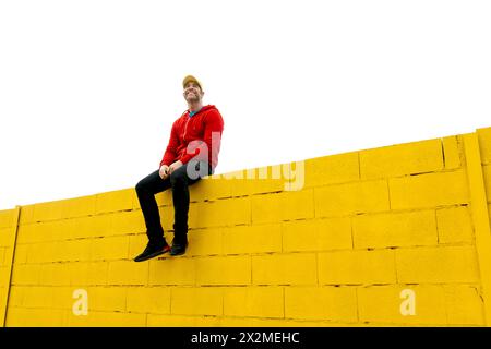 Un uomo gioioso in un abbigliamento casual si siede in cima a una parete di mattoni dipinti di giallo su uno sfondo bianco chiaro, trasuda un senso di libertà e felicità Foto Stock