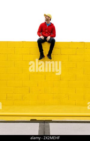 Un uomo con un abbigliamento casual siede su una parete gialla, creando un contrasto sorprendente con uno sfondo bianco Foto Stock