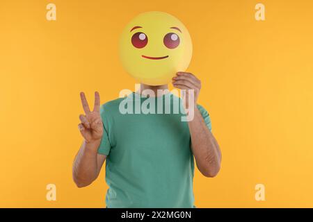 Uomo che copre il viso con emoticon sorridente e mostra un segno di pace su sfondo giallo Foto Stock