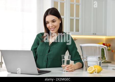 Donna con bicchiere d'acqua e caraffa del filtro vicino al notebook in cucina Foto Stock