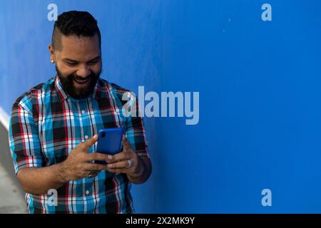 Uomo adulto (39) con barba e camicia a quadri, sorride mentre legge un messaggio di testo sul suo cellulare di colore blu, sfondo blu della parete. Copia spazio. Foto Stock