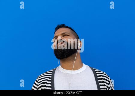Maschio adulto (39) con barba, piatto medio corto, ascolta la musica con le cuffie bianche. Guarda in alto dal lato destro. Vestito con maglione a righe, con Foto Stock