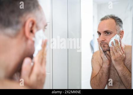 Un uomo applica schiuma da barba al viso in bagno Foto Stock