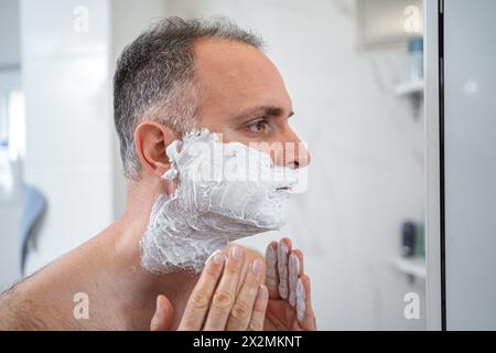 Un uomo applica schiuma da barba al viso in bagno Foto Stock