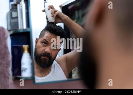 L'uomo latino (39) si taglia i capelli con un rasoio davanti a uno specchio. Concetto di barbiere. Foto Stock