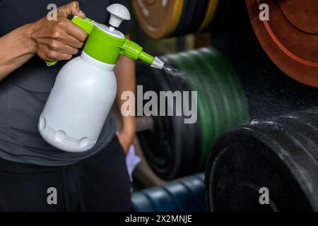 Primo piano delle mani di un uomo utilizzando una bottiglia di disinfettante per pulire i dischi di peso in palestra. Concetto di disinfezione, pulizia, antibatterico Foto Stock