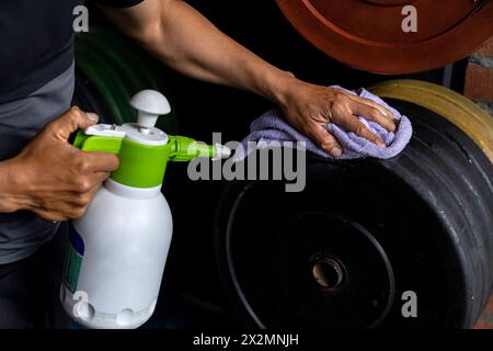 Primo piano delle mani di un uomo utilizzando una salvietta bagnata e una bottiglia di disinfettante per pulire i dischi di peso in palestra. Concetto di disinfezione, pulizia, anti-batteri Foto Stock