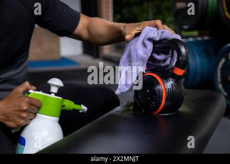 Primo piano delle mani di un uomo usando una salvietta bagnata e una bottiglia di disinfettante per pulire un kettlebell in palestra. Concetto di disinfezione, pulizia, anti-batteri Foto Stock