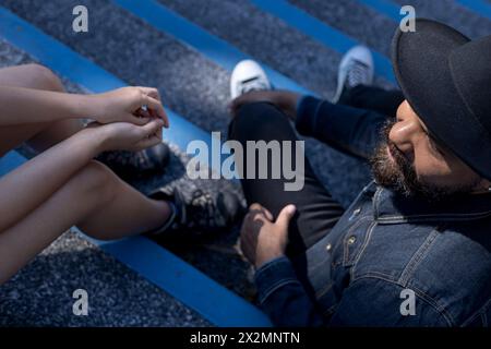 Vista dall'alto di un adulto con barba e cappello e di una giovane donna latinoamericana che ha una conversazione rilassata seduto sulle scale di una piazza. Riunione e Vaca Foto Stock