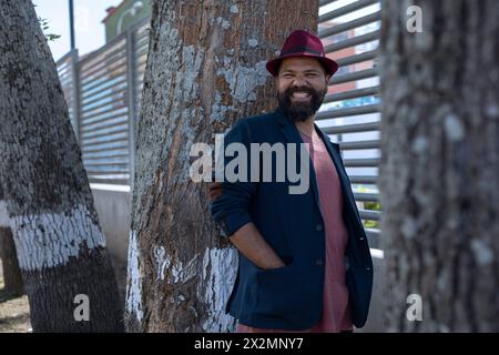 Colpo medio lungo di uomo bohémien latinoamericano sorridente con barba, cappello e giacca in piedi accanto ad alcuni alberi della città. Concetto di vacanza e portr Foto Stock