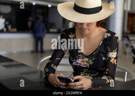 La giovane ragazza latinoamericana (22 anni) con cappello da cowboy sta mandando un messaggio dal suo cellulare. Concetto di tecnologia e vacanza Foto Stock