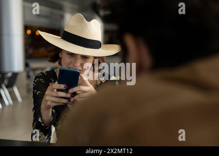 La giovane ragazza latinoamericana (22 anni) con cappello da cowboy sta mandando un messaggio dal suo cellulare. Concetto di tecnologia. Foto Stock