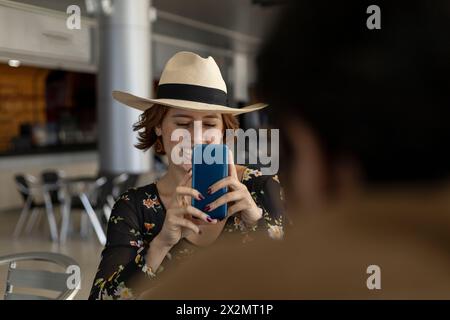 La giovane ragazza latinoamericana (22 anni) che indossa un cappello da cowboy sorride mentre scatta una foto con il suo cellulare. Concetto di tecnologia e vacanza Foto Stock