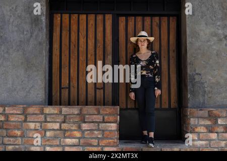 Giovane ragazza latino-americana (22) con cappello da cowboy posa davanti all'ingresso in legno sullo sfondo. Copia spazio. Concetto del modello Foto Stock