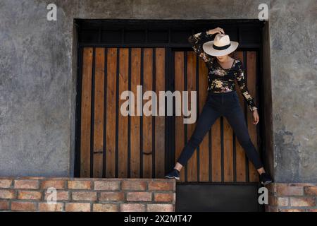 Giovane ragazza latino-americana (22) con cappello da cowboy posa davanti all'ingresso in legno sullo sfondo. Copia spazio. Concetto del modello Foto Stock